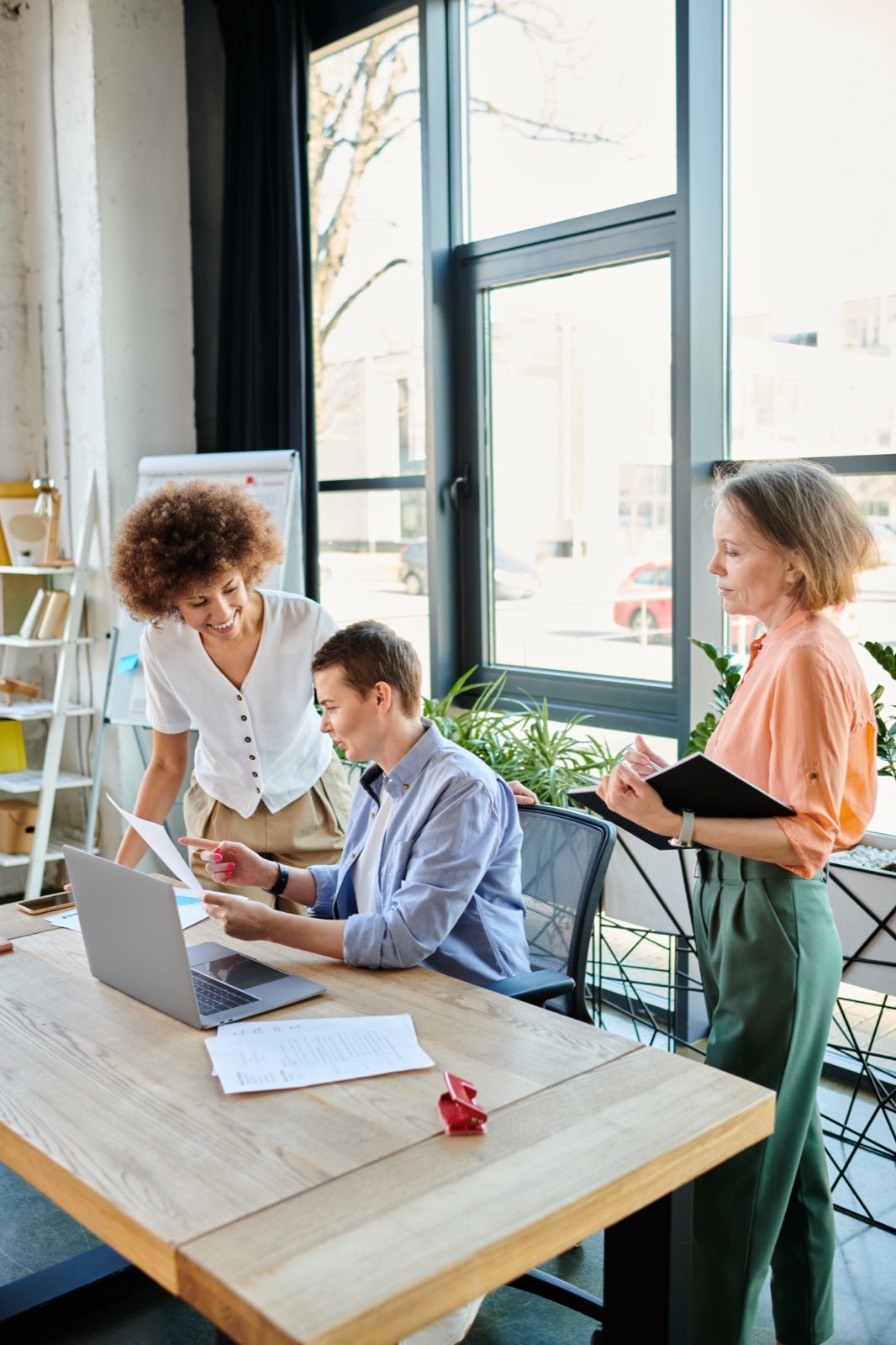 Three colleagues collaborating around a workspace in a bright office — one woman with curly hair leaning in and pointing at a printed page, another with short hair seated at a laptop, and a third standing to the right with an open notebook.