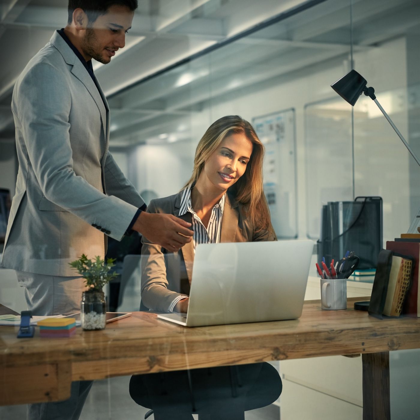 A man in a light gray blazer standing beside a seated colleague with long blonde hair, both smiling at her laptop screen while reviewing a project together in a modern glass-walled office.