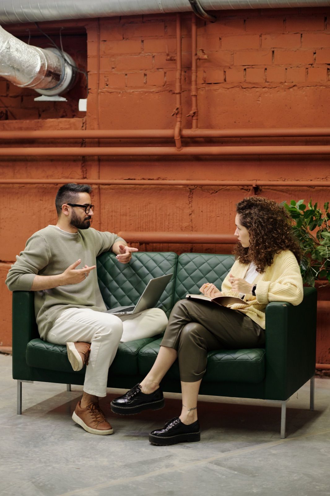 Two colleagues mid-conversation on a dark green leather sofa in a warm brick-walled loft — a man in an olive sweater gesturing to explain a point while a woman in a cream cardigan listens, notebook and laptop open on her lap.